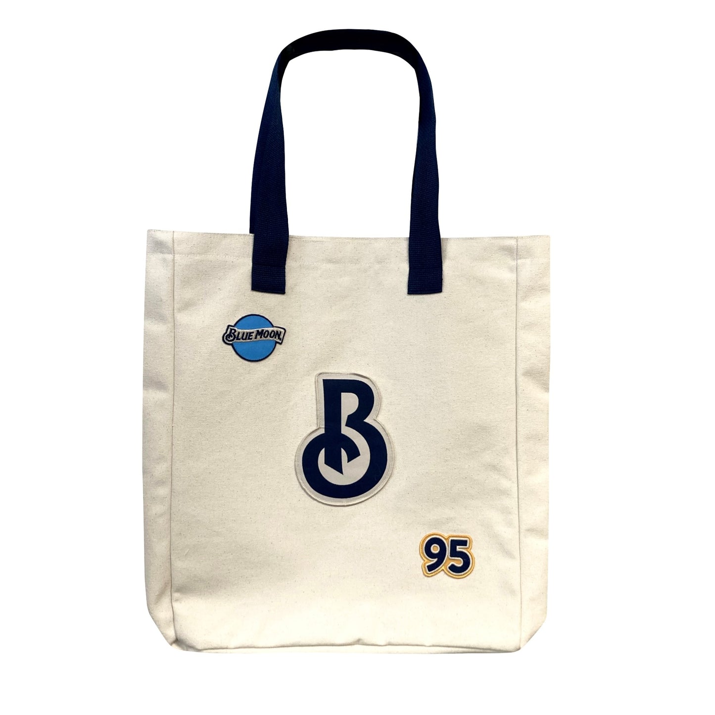 Cream canvas tote bag with navy handles, a small Blue Moon logo patch on the upper left, a large stylized “B” patch in the center, and a “95” patch in the lower right.