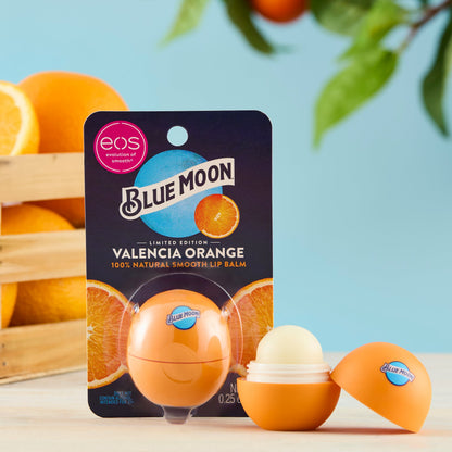 Orange eos-style lip balm container with the Blue Moon logo, shown open next to its Valencia Orange package, with whole oranges and leaves in the background.