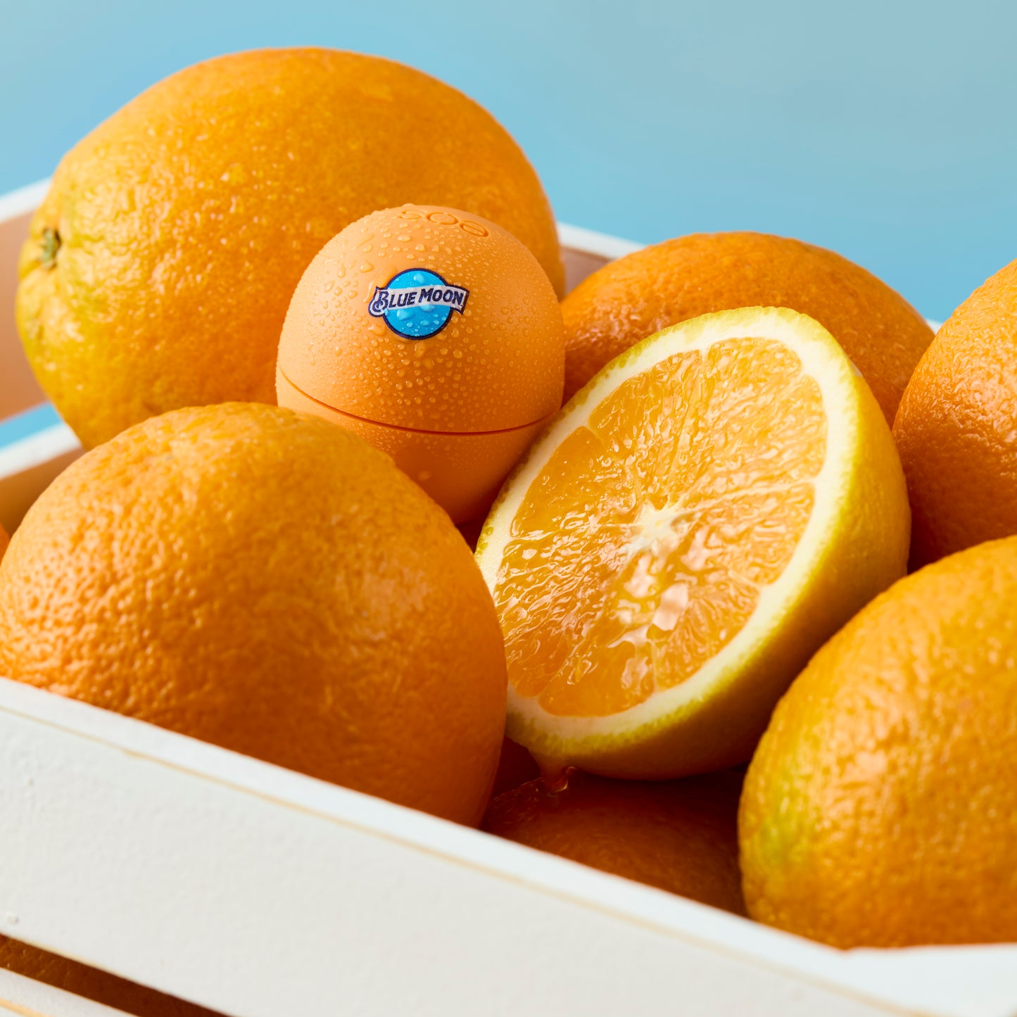 Orange Blue Moon–branded spherical lip balm resting among whole oranges and a sliced orange in a white crate.