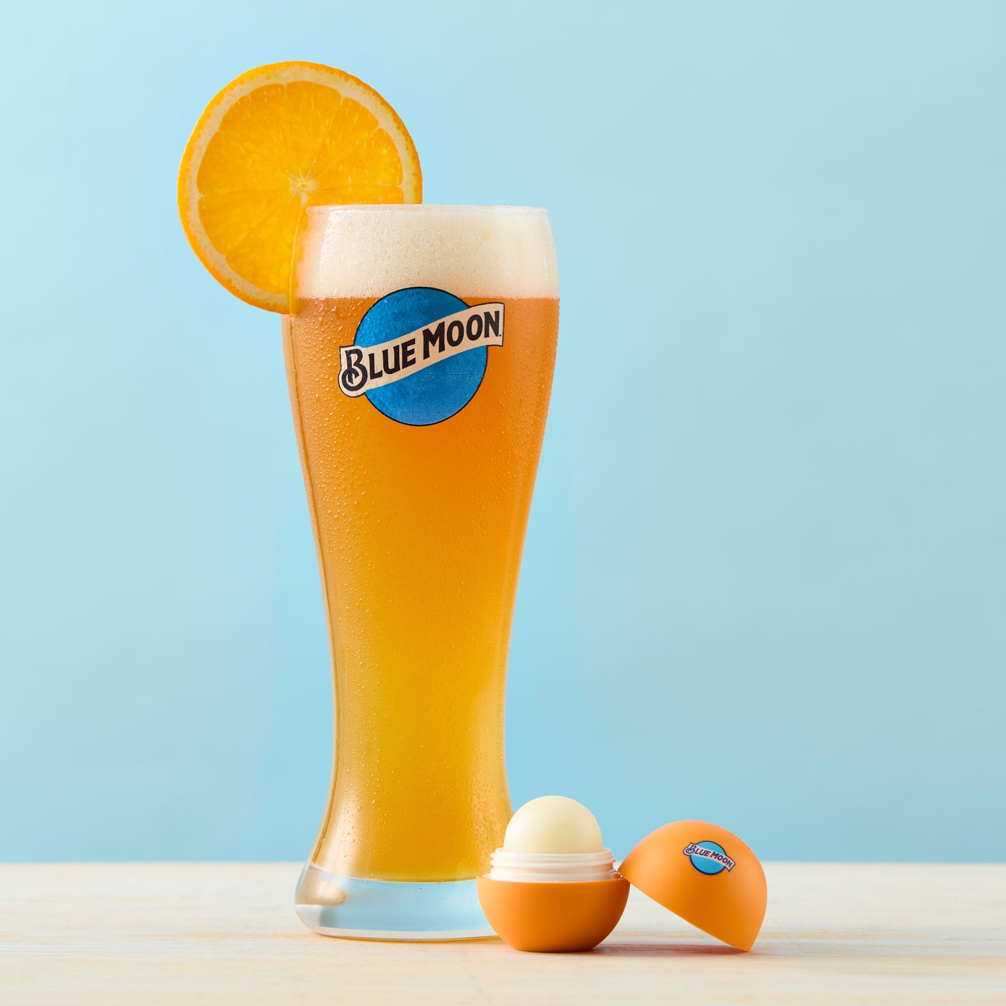 Open orange Blue Moon–branded lip balm container, beside an tall Blue Moon beer glass with foam and an orange slice garnish.