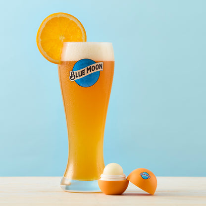Open orange Blue Moon–branded lip balm container, beside an tall Blue Moon beer glass with foam and an orange slice garnish.