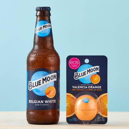 Valencia Orange Blue Moon–branded eos-style lip balm package next to a brown Blue Moon Belgian White beer bottle 